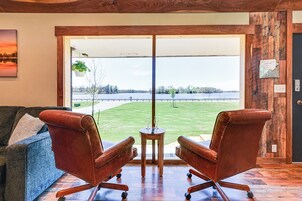 Una habitación con un ventanal amplio, dos sillones de cuero, una mesita de madera con dos copas de vino y vistas a un lago.