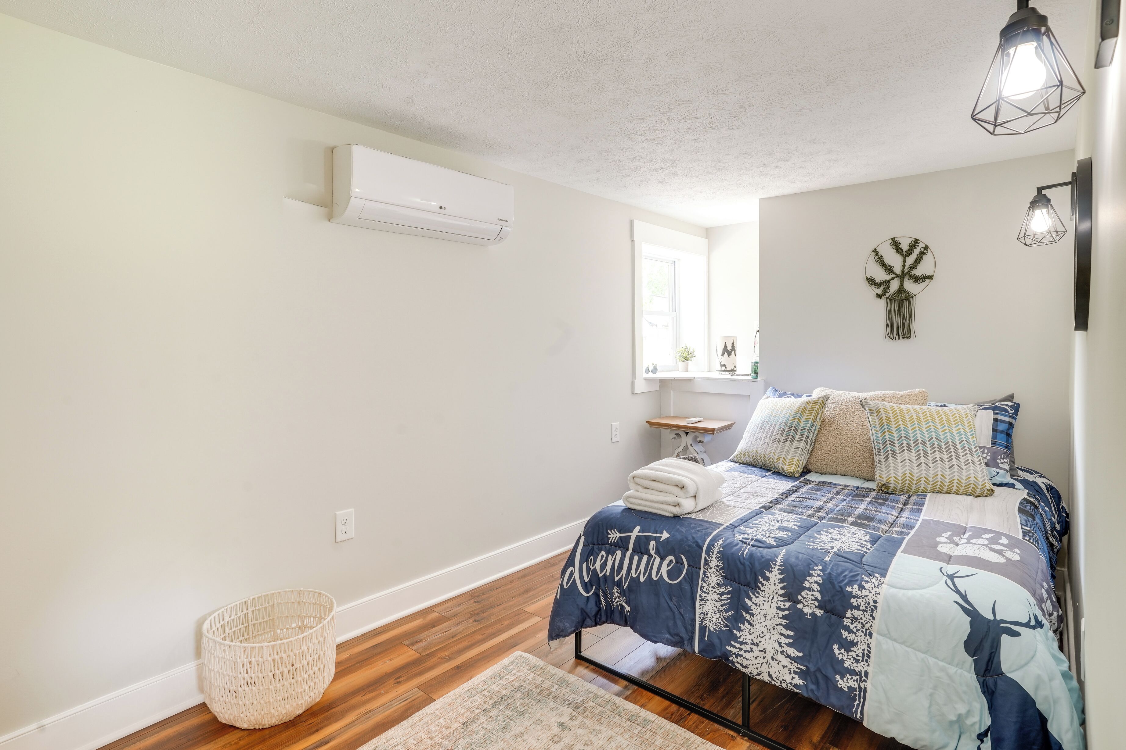 A bedroom with a bed, a window, an air conditioner, a wall-mounted shelf, and a basket.