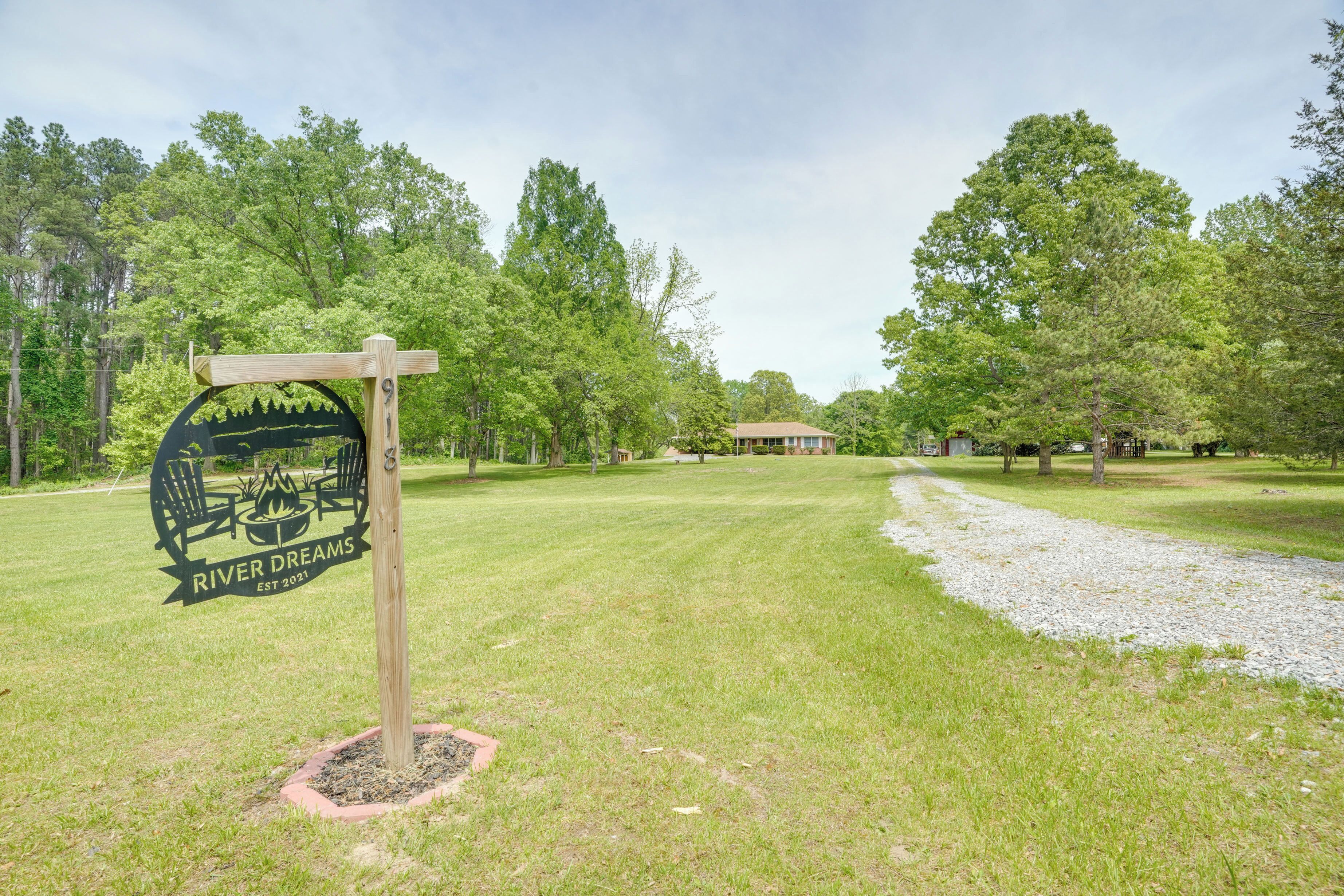 A sign for River Dreams with a logo of a campfire and chairs, surrounded by a grassy area with trees and a gravel path.