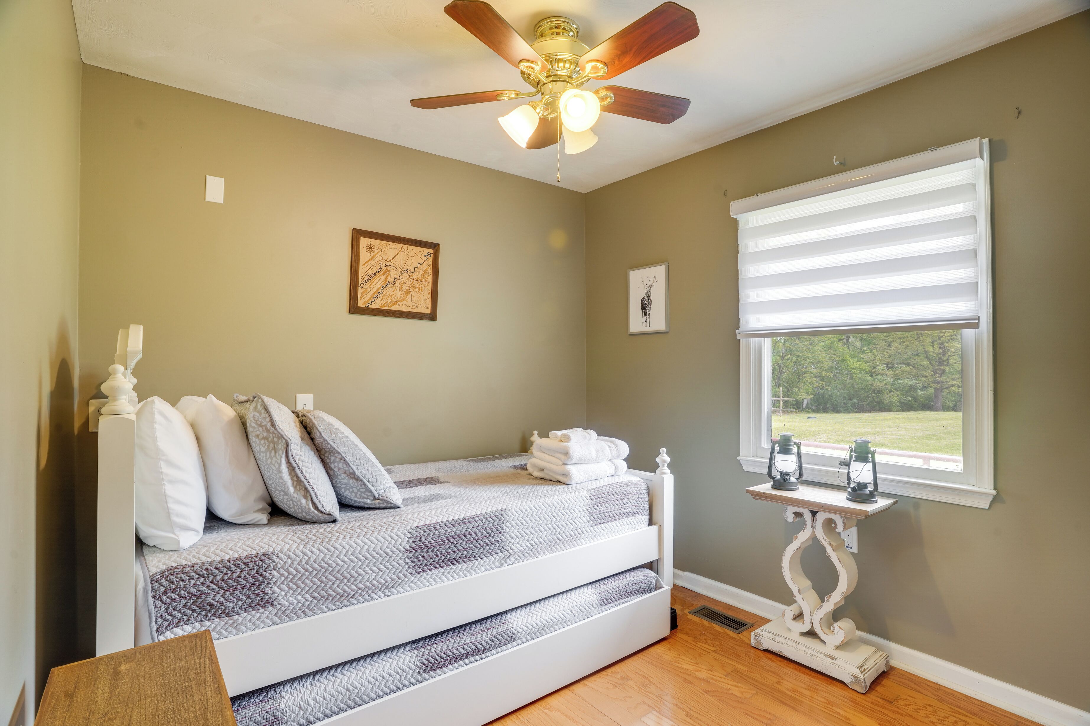A bedroom with a bed, a ceiling fan, a window with blinds, and a small table with two lanterns.
