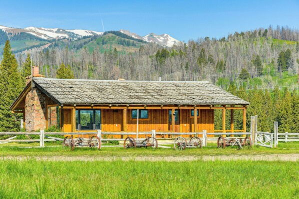 A rustic cabin with a stone chimney, wooden walls, and a porch with vintage wagon wheels. - Rustic Bondurant Cabin: Fish, Hike, & Bike! (Bondurant)
