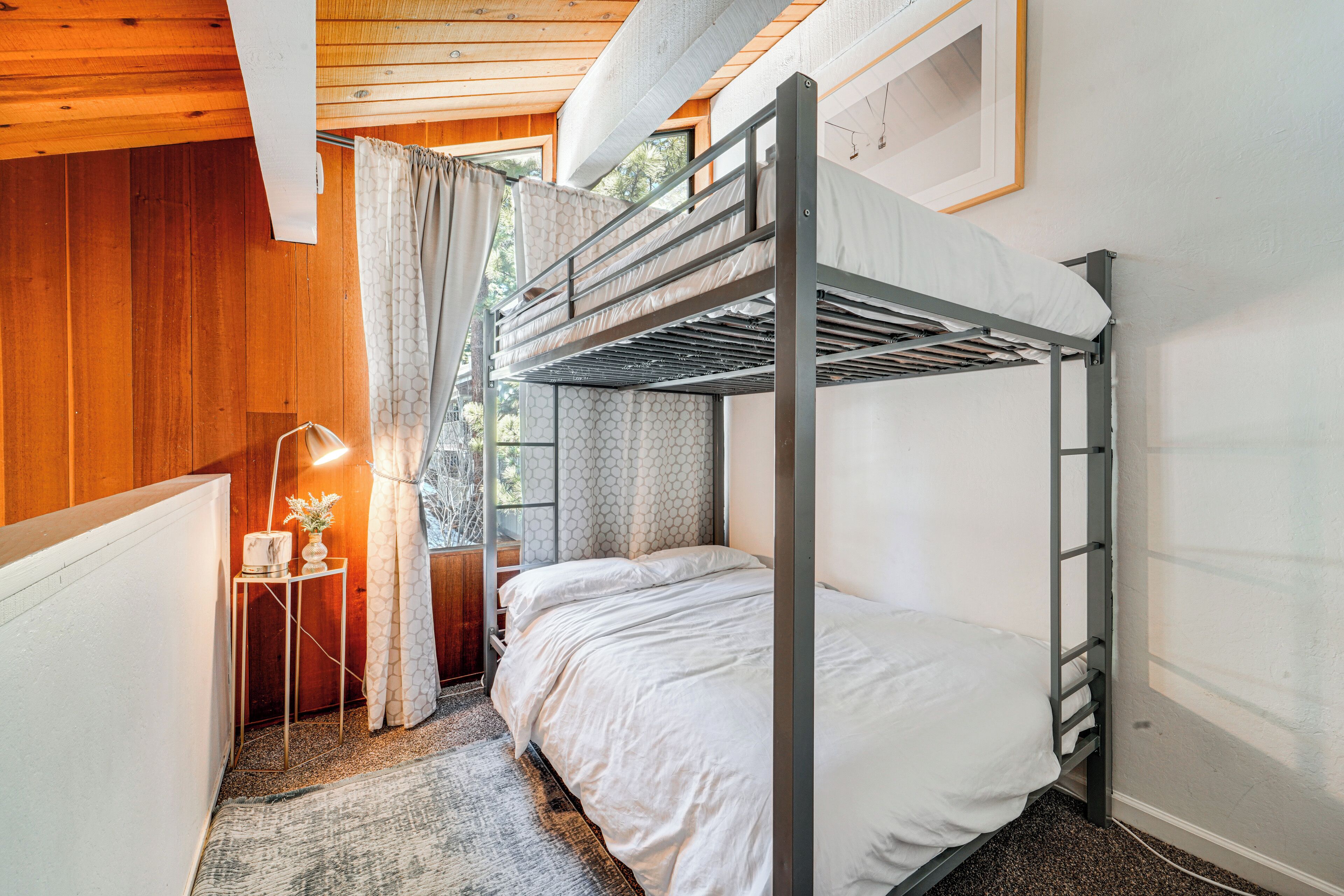 Dormitorio con literas, techo de madera, una mesita con lámpara y ventana con cortinas.
