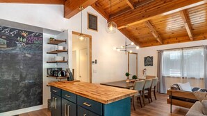 A modern kitchen with wooden cabinets, a central island, and pendant lighting. There is a dining area with a table and chairs, and a living space with a sofa and a window.