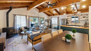 A spacious living area with a wooden ceiling, a fireplace, a dining table with chairs, and a kitchen area with a refrigerator and shelves.