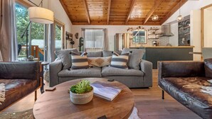 A cozy living room with a wooden ceiling, a grey sofa with patterned cushions, a round coffee table with a plant, and a leather couch.