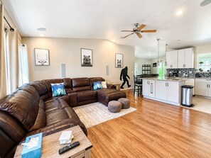 A modern living room with a leather sectional sofa, a wooden coffee table, and a kitchen area with white cabinets and a stainless steel trash can. - Big Foot Basecamp - Dogs / Hot Tub / Fire Pit (South Lake Tahoe)