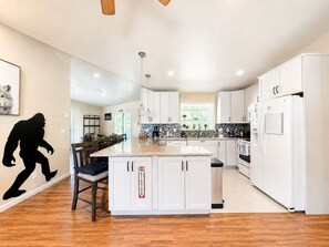 A modern kitchen with white cabinets, a central island, and a dining area with a chair. - Big Foot Basecamp - Dogs / Hot Tub / Fire Pit (South Lake Tahoe)