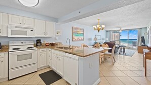 A spacious kitchen with white cabinets, a built-in microwave, and a stove. There is a dining area with a table and chairs, and a view of the ocean through large windows. - Pelican Beach 1115 2 Bedroom Condo (Destin)