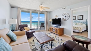 A spacious living room with a balcony overlooking the ocean, a television, and a bed with a floral bedspread. - Pelican Beach 1115 2 Bedroom Condo (Destin)