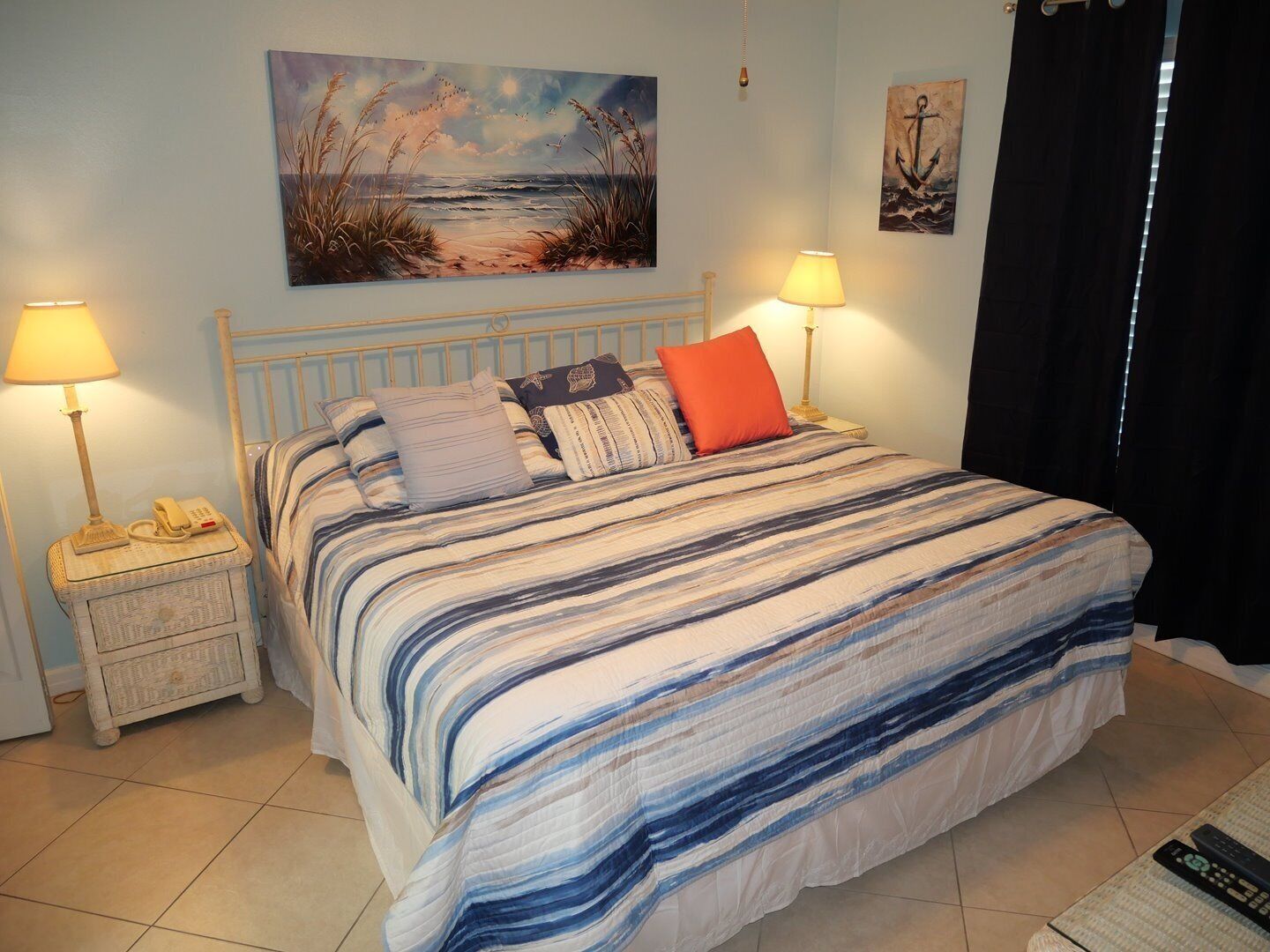 A bedroom with a bed, two bedside tables with lamps, a painting on the wall, and a curtain.