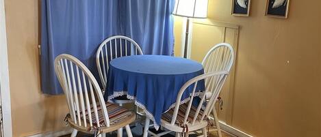 A dining area with a round table covered with a blue tablecloth, surrounded by four white chairs with striped cushions. A blue curtain with a floral pattern is partially drawn, and a sign above reads "What Happens At The Cottage Stays At The Cottage."