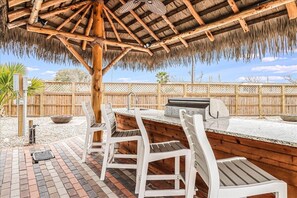 An outdoor patio with a wooden table and chairs, a grill, and a thatched roof overhead. - Margarita Time Sfth1 1 Bedroom Home by RedAwning (Port Aransas)
