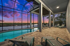 Piscina interna com vista para um pôr do sol sobre a água, cercada por espreguiçadeiras e um terraço coberto.