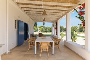 A covered patio with a white table and wicker chairs, a blue door, and a view of greenery. - Casa Blue con piscina by Wonderful Italy (Ostuni)