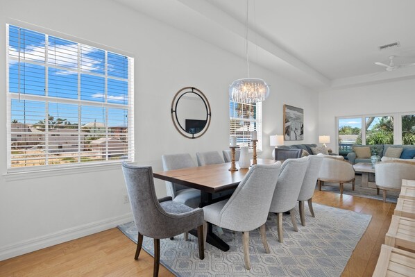 A modern living room with a dining area, large windows, and a chandelier.