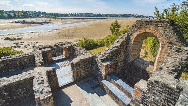 Een oud stenen gebouw met een grote boog, een stenen trap en een uitzicht op een strand en een waterlichaam op de achtergrond.
