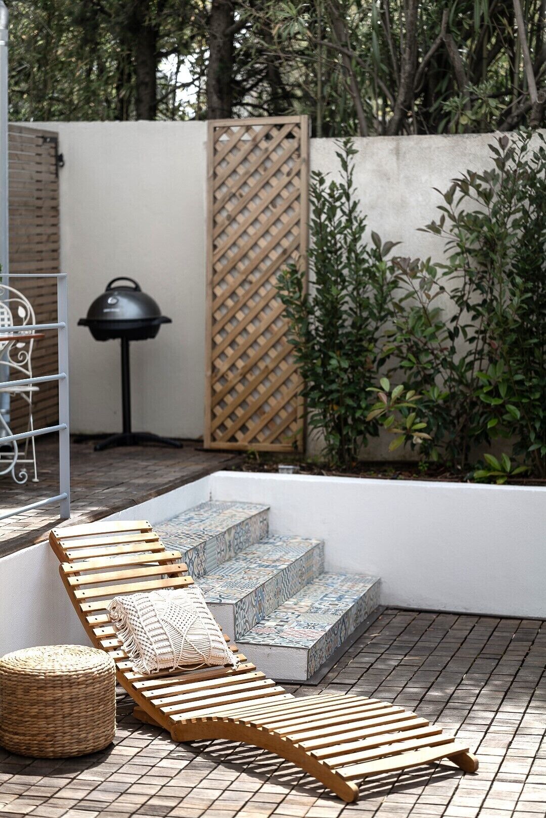 A patio with wooden loungers, a woven ottoman, and a patterned stone staircase.