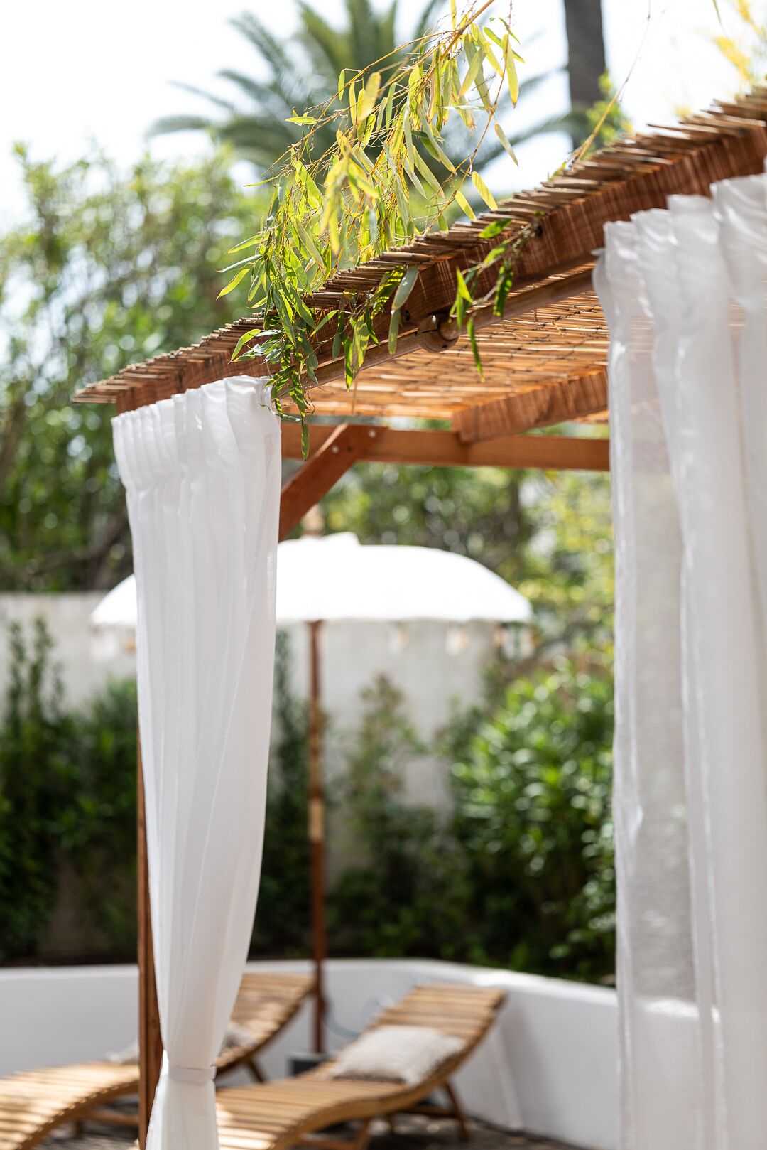 Una pérgola de madera con cortinas blancas, un paraguas blanco y una silla de descanso.
