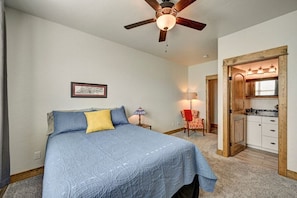 A bedroom with a bed, a ceiling fan, a nightstand, a lamp, a chair, and a kitchen area visible through an open door.