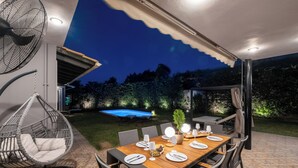 An outdoor dining area with a wooden table set for four, surrounded by chairs. There is a hanging chair and a pool in the background.