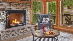 A stone fireplace with a fire, a grey armchair with a deer pillow, and a round table with a bottle, wine glass, and snacks.