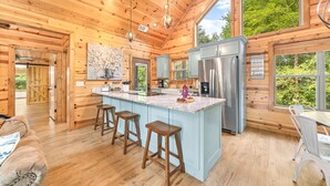 A wooden cabin interior with a kitchen island, bar stools, a refrigerator, and a large window.