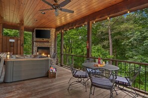 A covered outdoor deck with a stone fireplace, a hot tub, and a table with chairs.