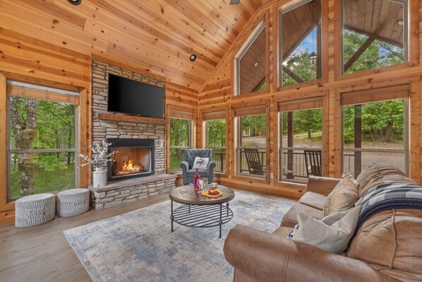A log cabin interior with a stone fireplace, wooden walls, large windows, and a furnished living area.