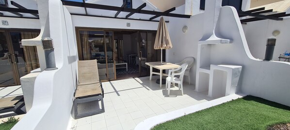 A modern outdoor patio area with a table and chairs, a small wooden bench, and a glass door leading to an interior space.
