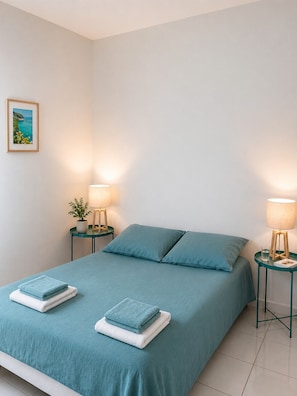 Un letto rifatto con lenzuola color petrolio e due cuscini, affiancato da due comodini con lampade, una piccola pianta in vaso e un quadro appeso al muro.