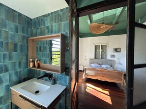 A bathroom with a modern sink and mirror, a wooden shelf with toiletries, and a view of a bedroom with a bed and window. - Louisette à la Rivière  (La Rivière St Louis)
