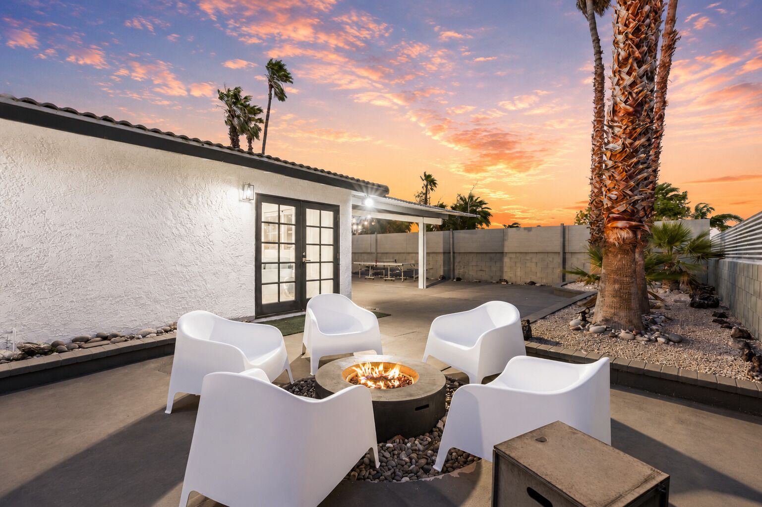 An outdoor patio with white chairs, a fire pit, and a view of the sunset.