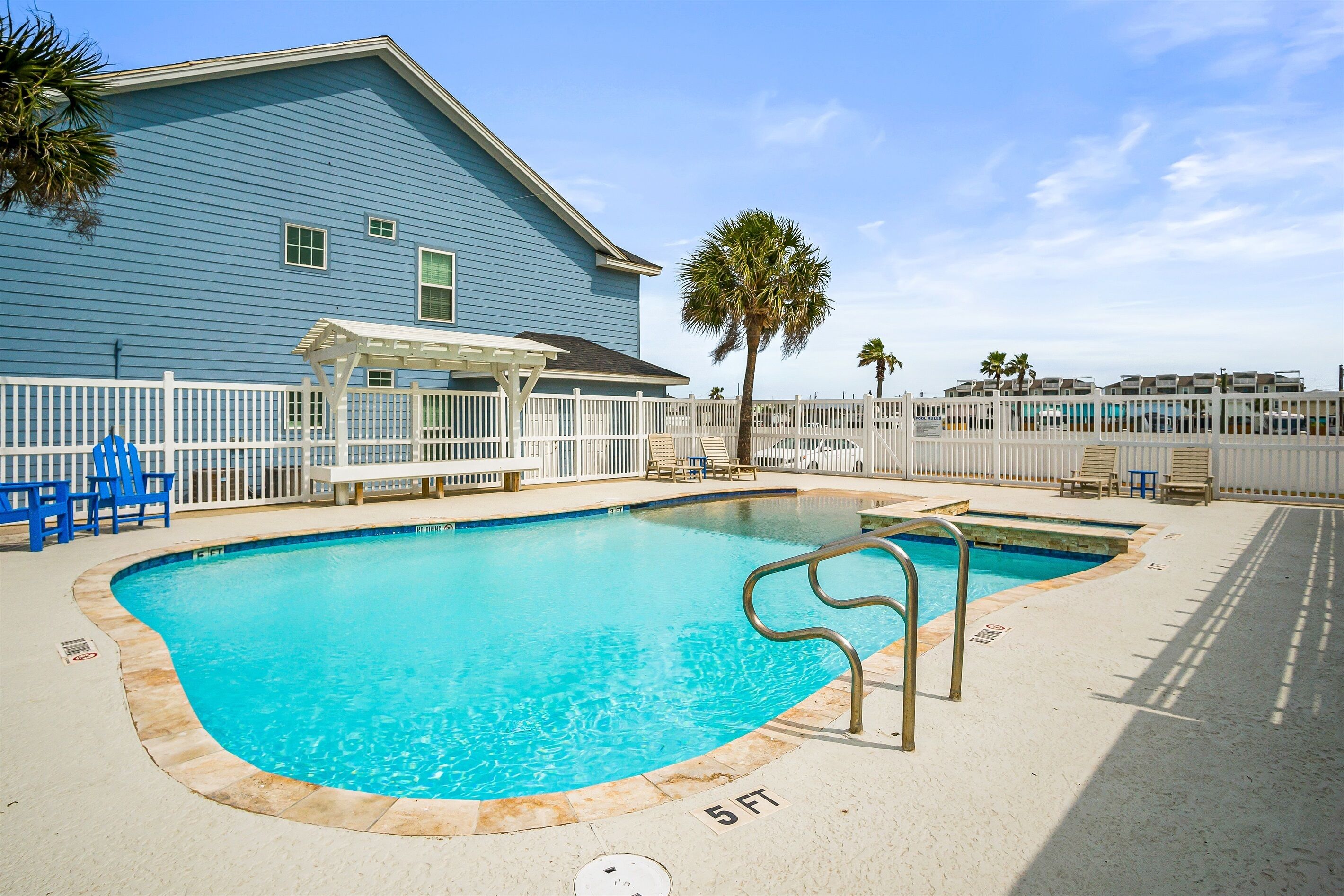 A swimming pool with a white fence, a covered seating area, and a blue building in the background.