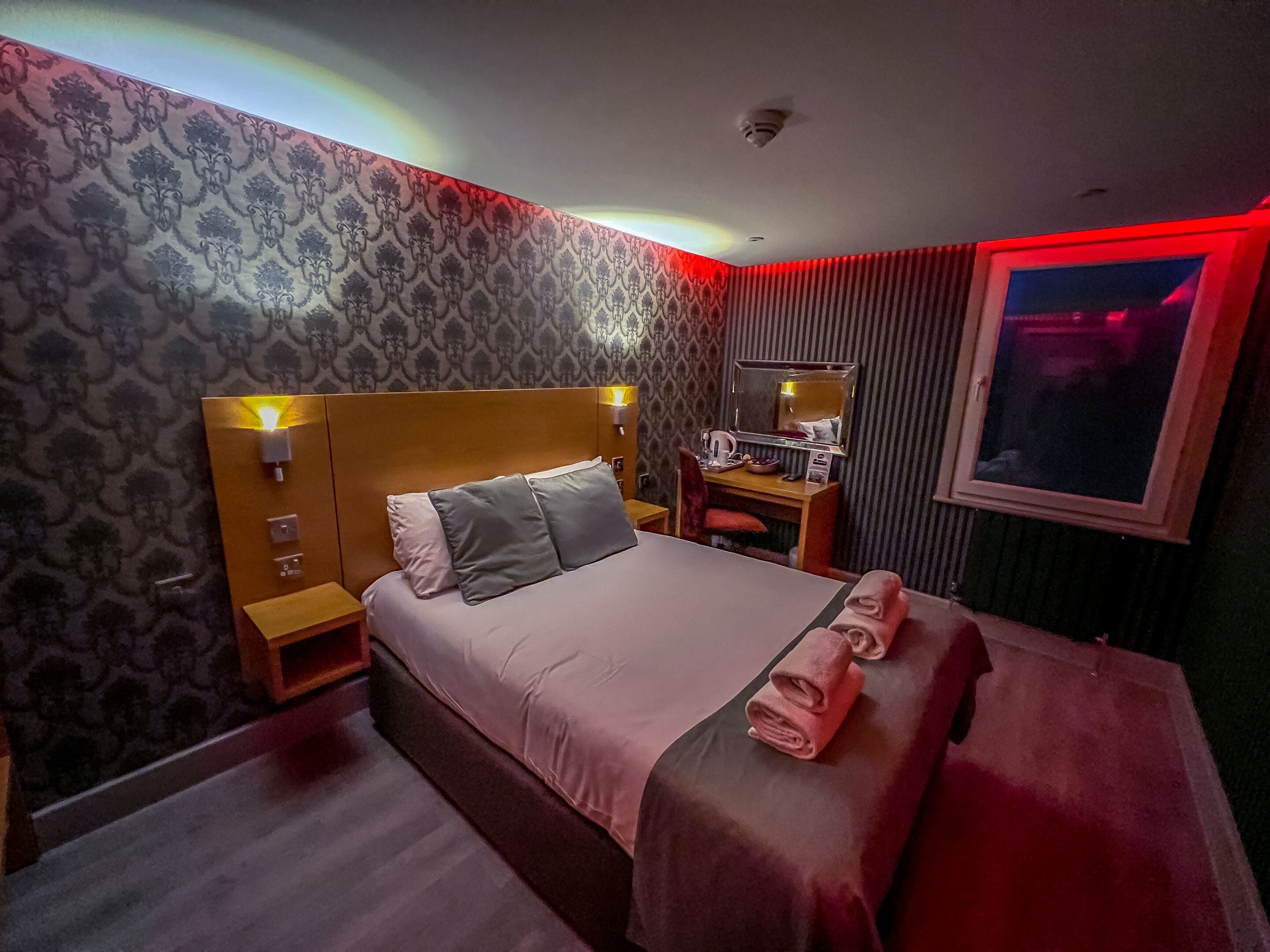 A hotel room with a large bed, a bedside table, a nightstand, a desk with a mirror, and a window with red lighting.
