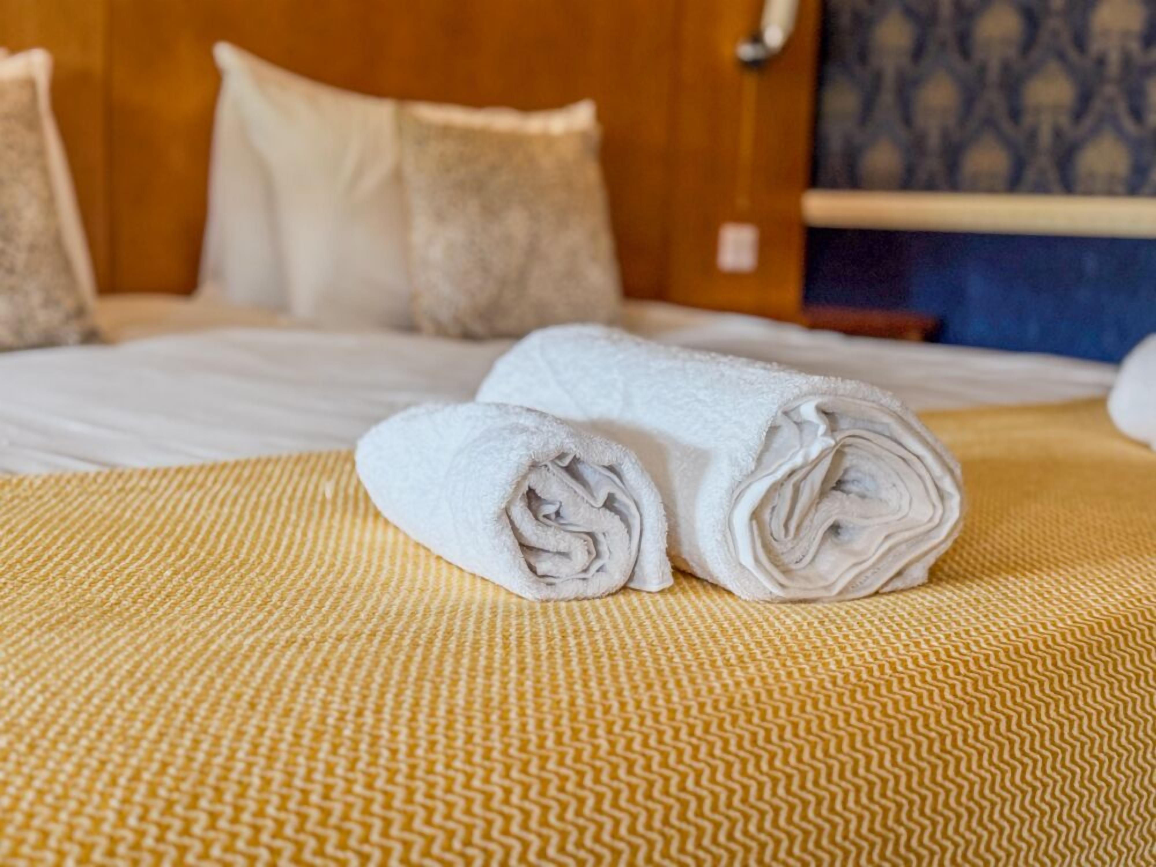 Una cama bien hecha con toallas blancas enrolladas y una colcha amarilla estampada.