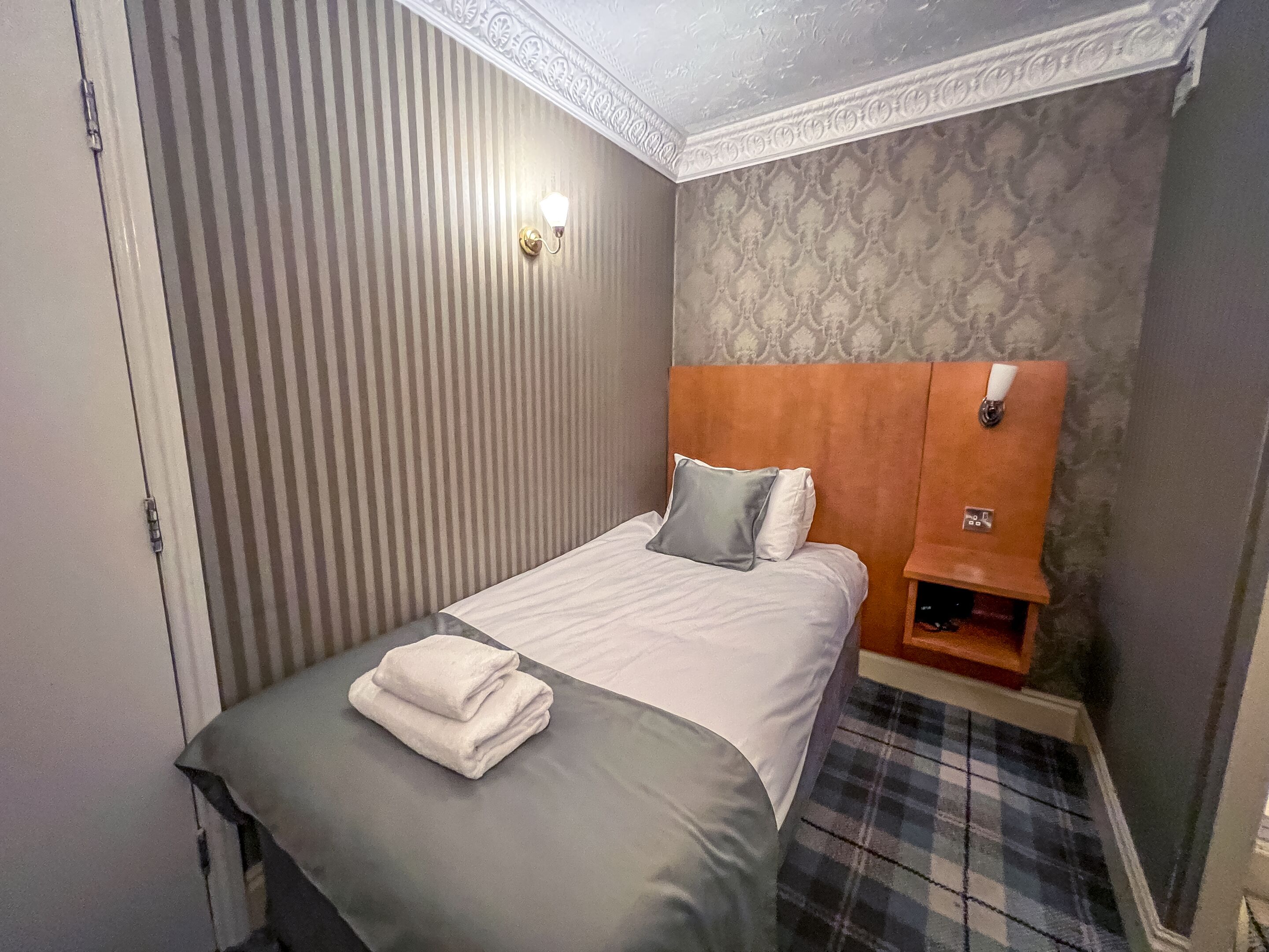 A neatly made bed with a folded towel, a headboard with a small shelf, and a striped wall.