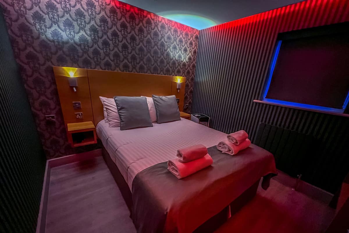A modern hotel room with a large bed, a headboard with built-in lighting, a flat-screen TV, and a red carpeted floor.