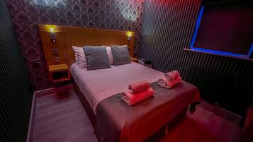 Una habitación de hotel moderna con una cama grande, cabecera con iluminación integrada, televisor de pantalla plana y piso cubierto de alfombra roja.