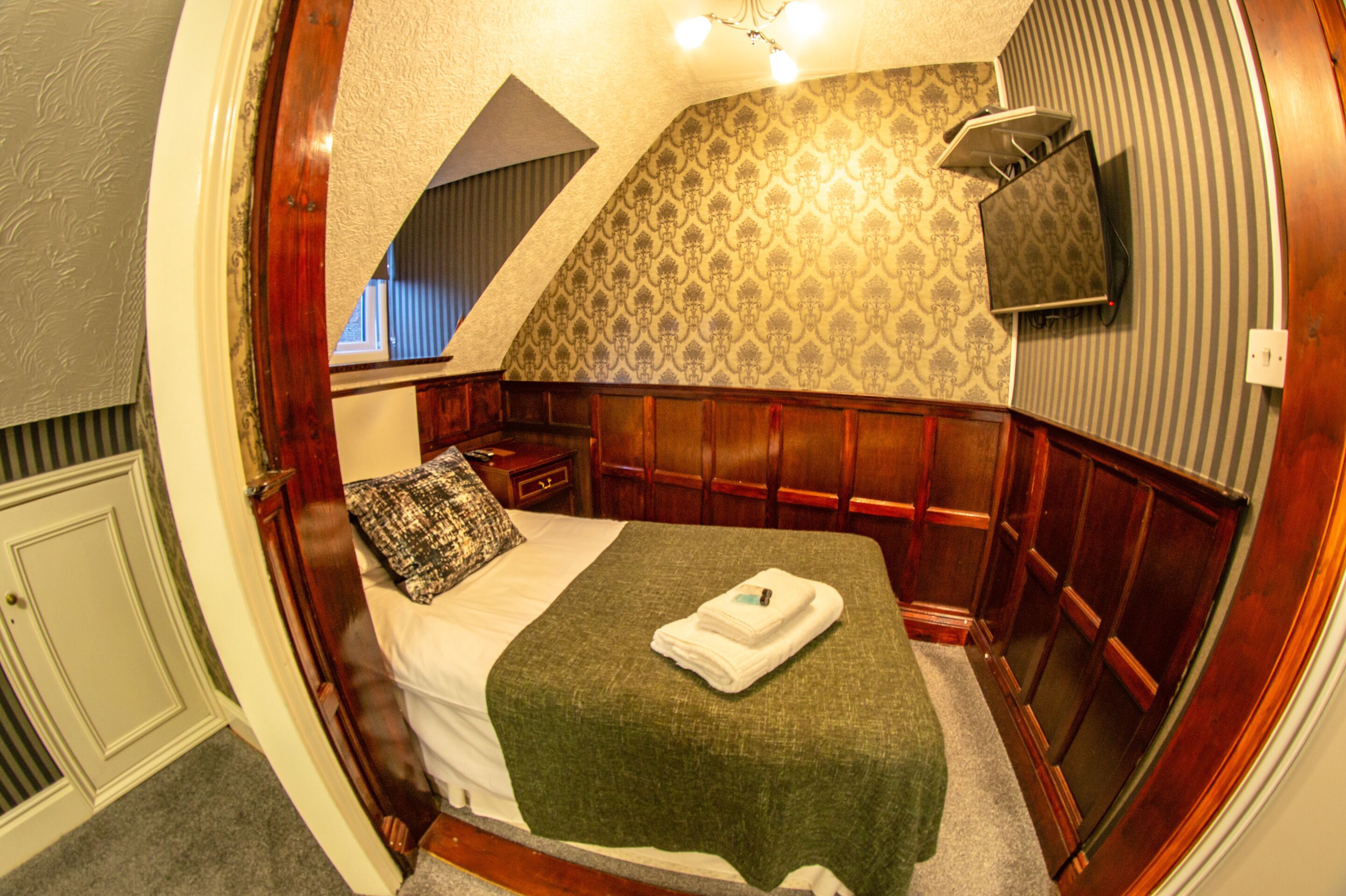 A bedroom with a sloped ceiling, a bed with a green blanket, a wooden headboard, a nightstand, and a wall-mounted TV.