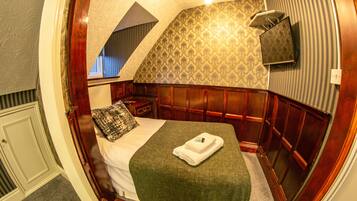 A bedroom with a sloped ceiling, a bed with a green blanket, a wooden headboard, a nightstand, and a wall-mounted TV.
