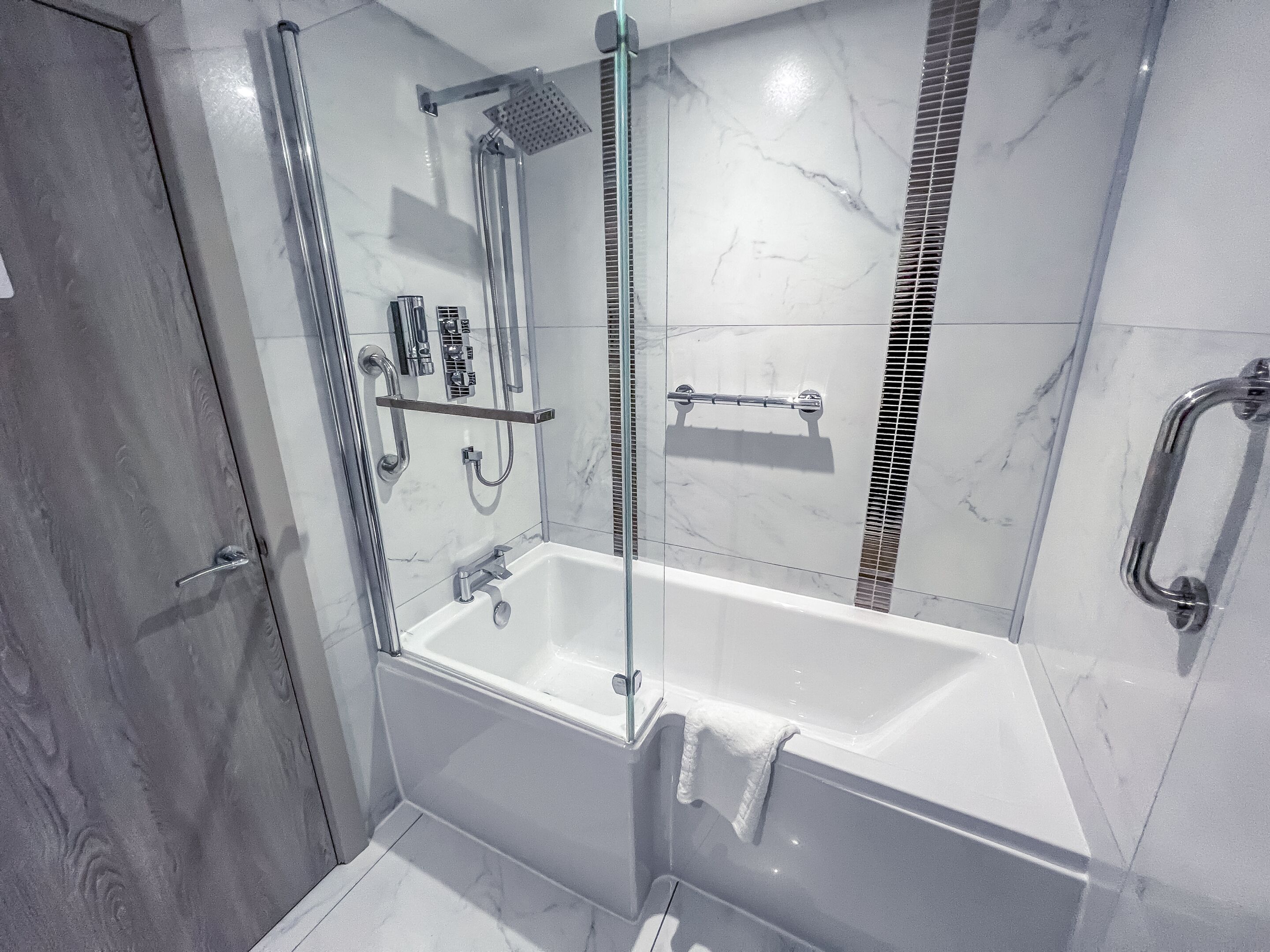 A modern bathroom with a glass-enclosed shower, a white bathtub, and a handrail.