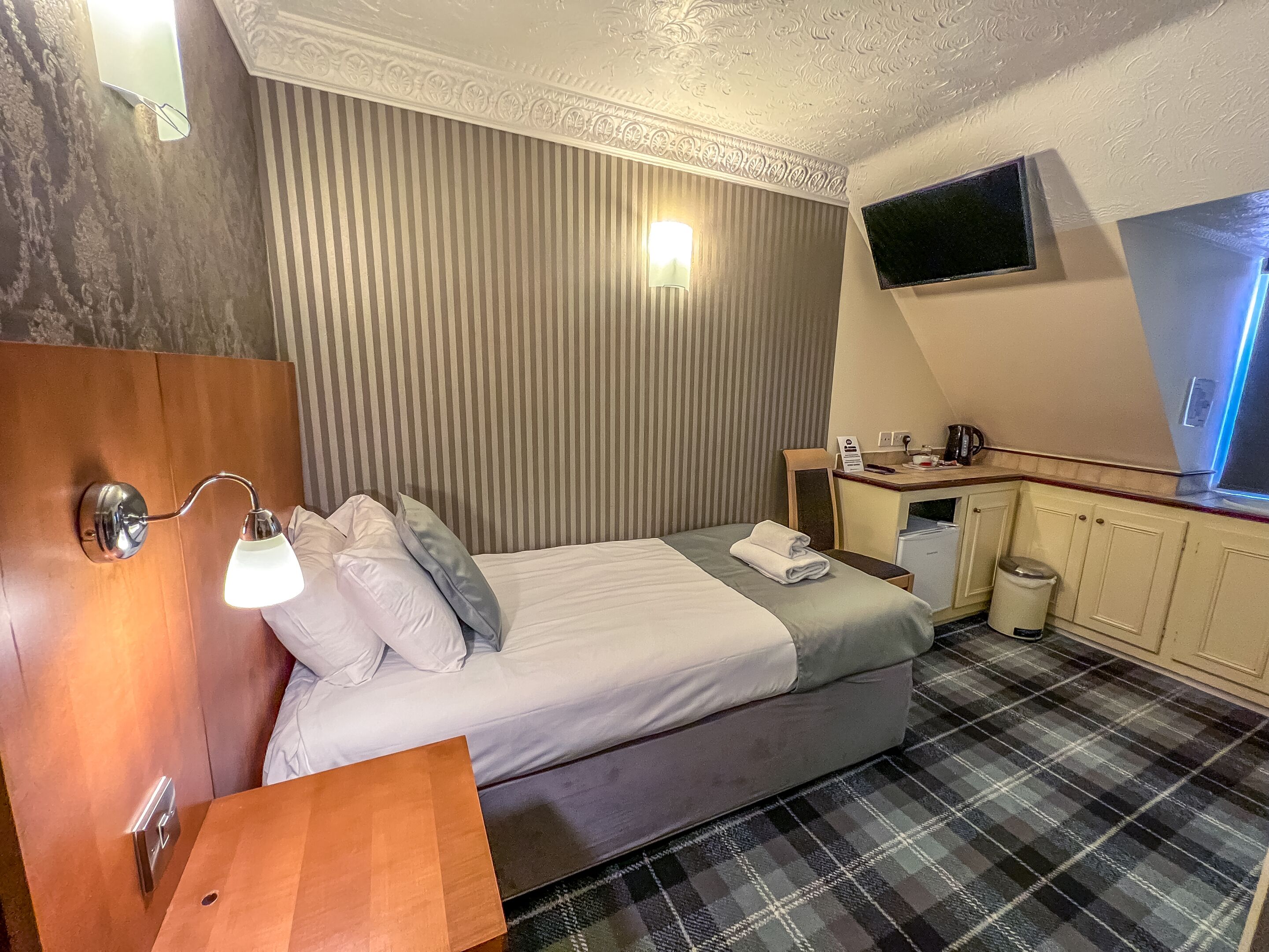 A hotel room with a bed, bedside lamp, a small wooden table, a TV mounted on the wall, and a kitchenette area.