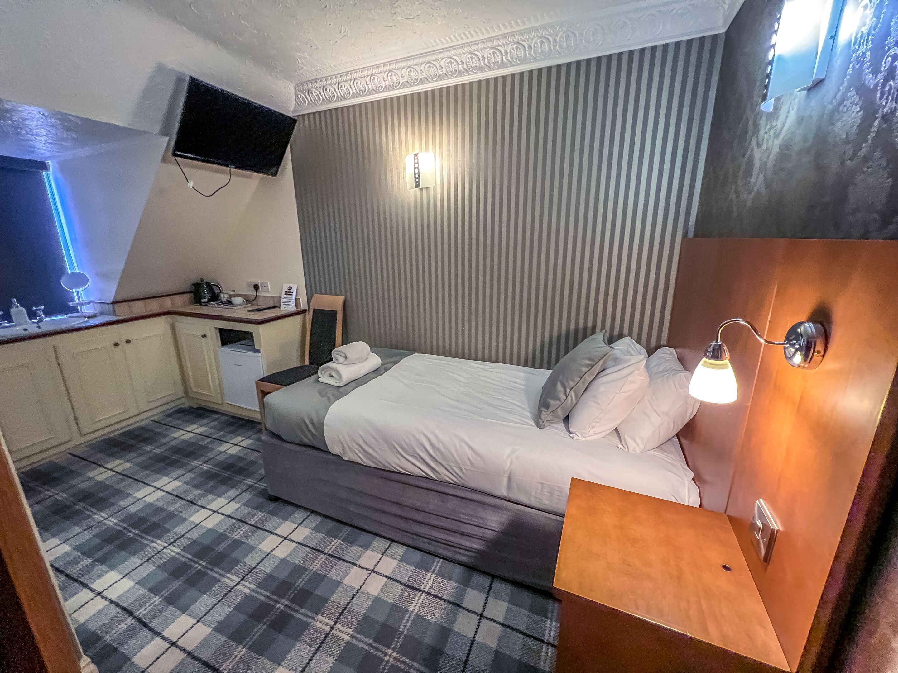 Ein Hotelzimmer mit Bett, Nachttisch, kleinem Schreibtisch, Stuhl, einem an der Wand befestigten Flachbildfernseher und einer Rezeption.