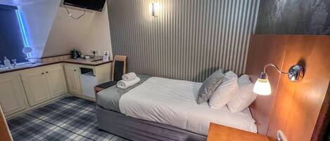 A hotel room with a bed, a bedside table, a small desk, a chair, a flat-screen TV mounted on the wall, and a check-in counter.