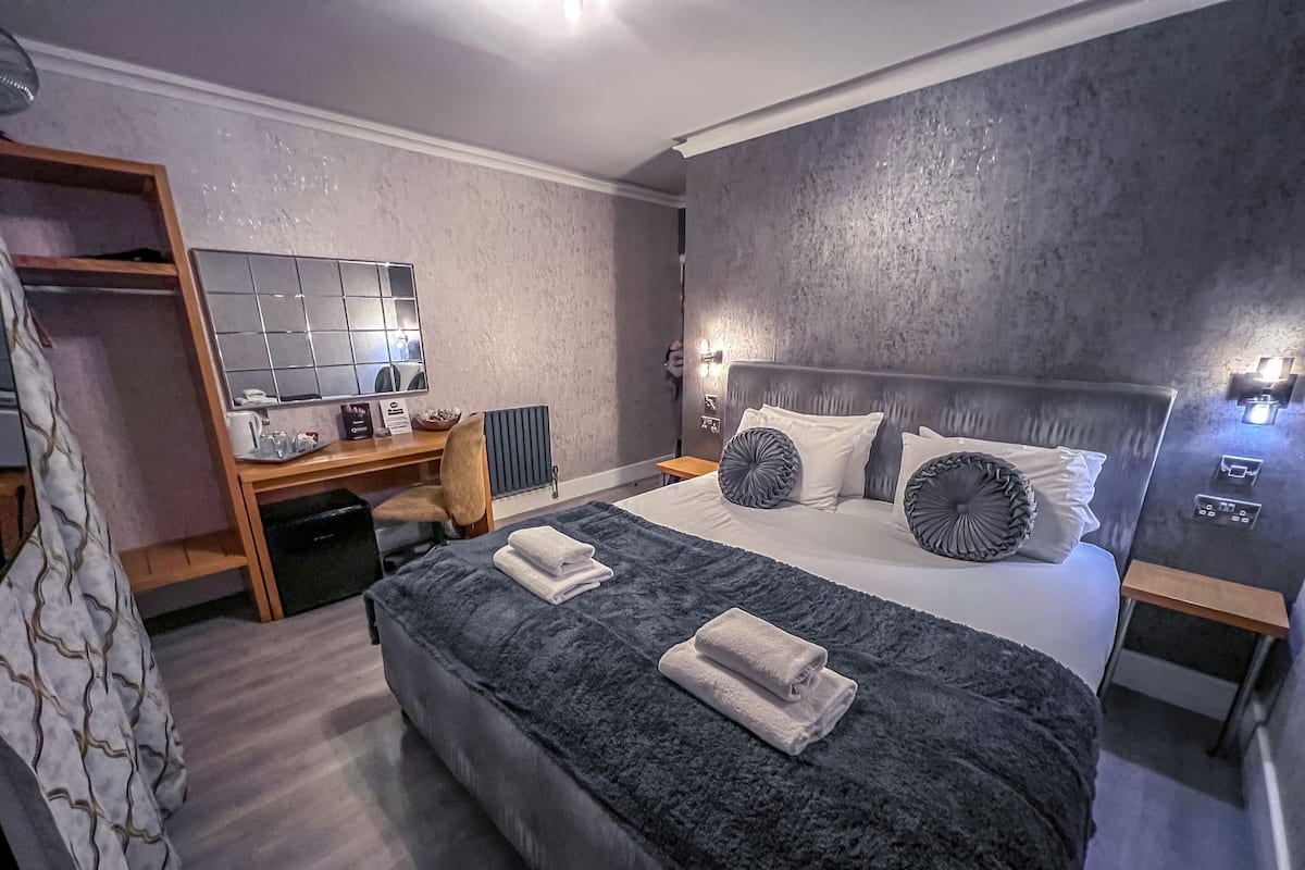 A modern hotel room with a large bed, a desk with a computer, a chair, a wardrobe, and a bathroom.