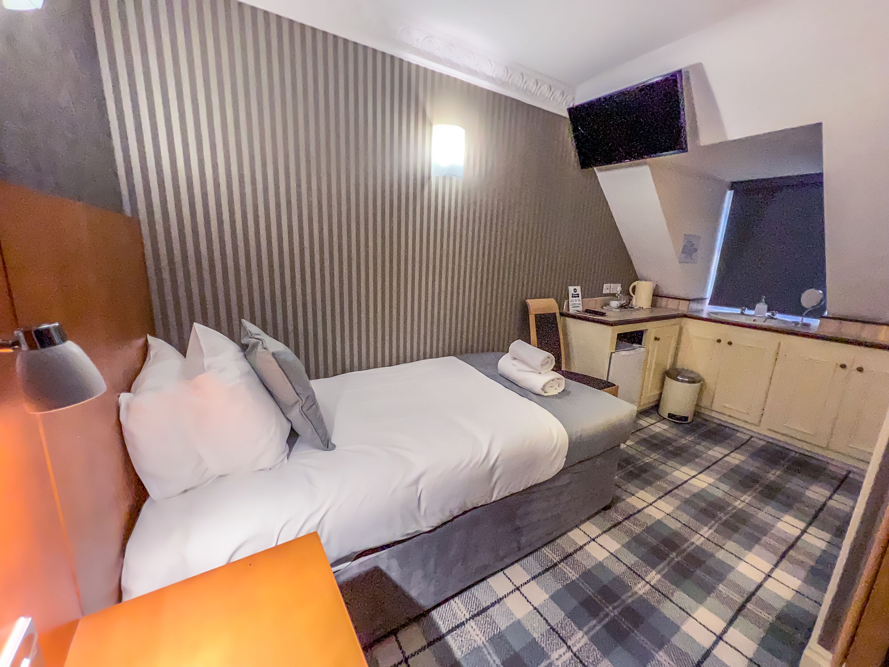 A hotel room with a bed, a bedside table with a lamp, a small kitchenette, and a check-in counter.