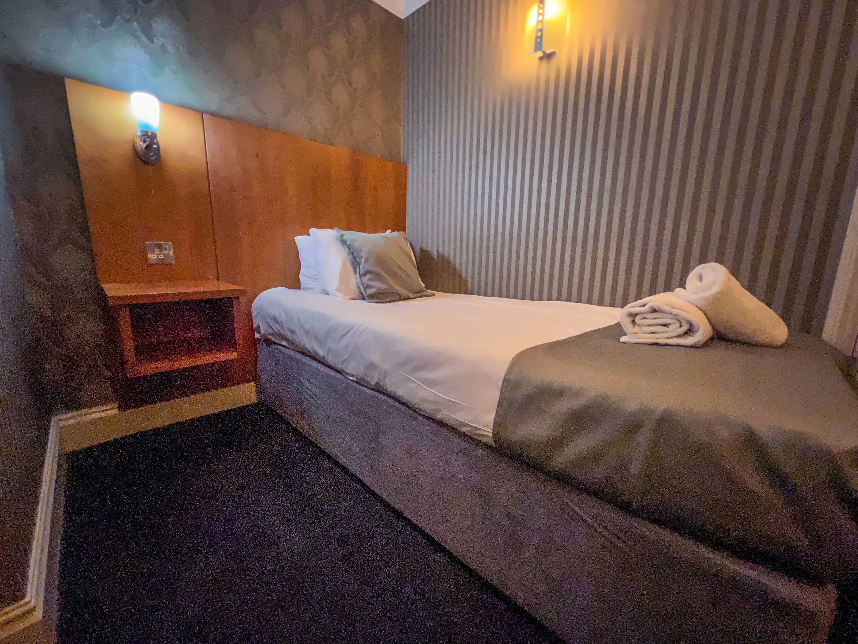 A hotel room with a bed, a nightstand, a wall-mounted lamp, and a striped curtain.
