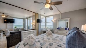A bedroom with a bed, a ceiling fan, a TV, a desk, a chair, and a view of the ocean.
