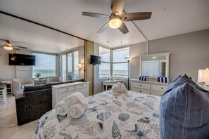 A bedroom with a bed, a ceiling fan, a TV, a desk, a chair, and a view of the ocean.