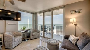 A living room with a sofa, armchair, ottoman, and a flat-screen TV. There is a balcony with a view of the ocean.